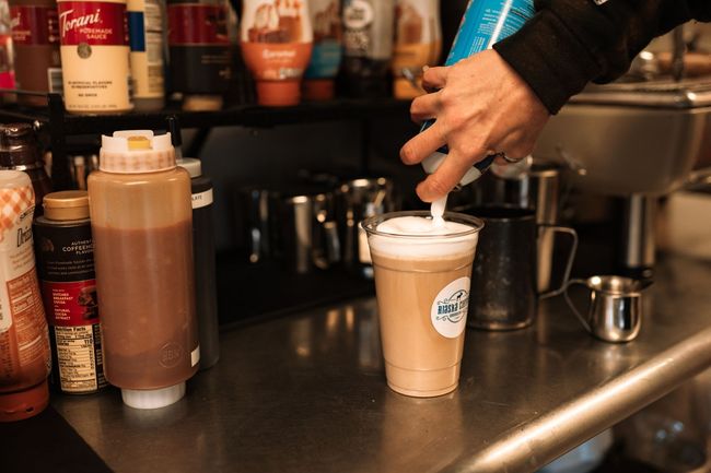 A person uses a whipped cream canister to top a coffee drink in a plastic cup inside a café workspace.