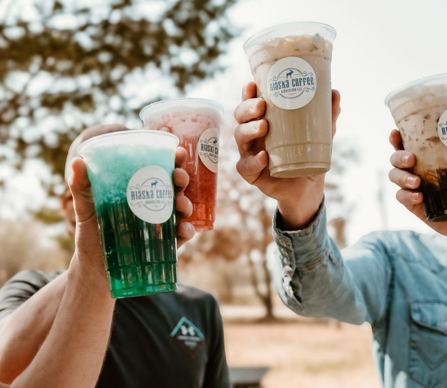 Three hands hold clear plastic cups of colorful iced drinks up for a toast outdoors against a blurred tree background.