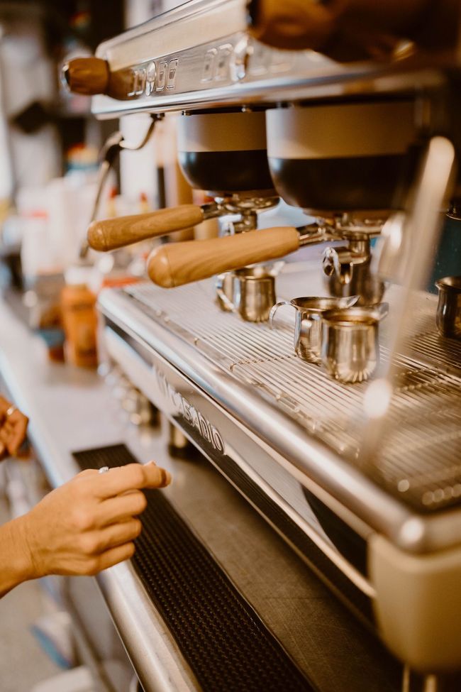 A person's hand reaches toward a professional espresso machine with wooden handles in a cafe setting.