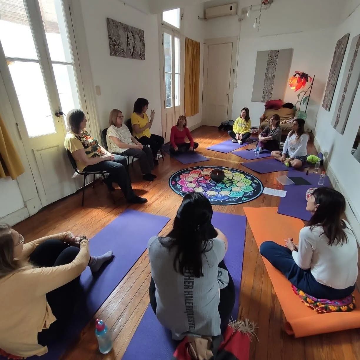 Grupo sentado en colchonetas de yoga en una habitación iluminada por el sol, con un mandala colorido en el centro.