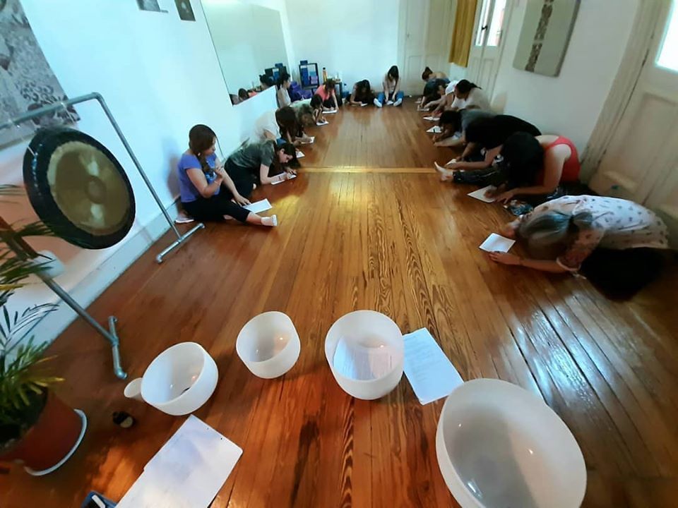 Personas sentadas sobre un suelo de madera, estirándose. Un gong y cuencos sonoros se ven en primer plano, en una sala soleada.