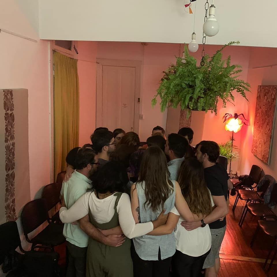 Un grupo de personas abrazándose en una habitación con un helecho colgante y una iluminación suave.