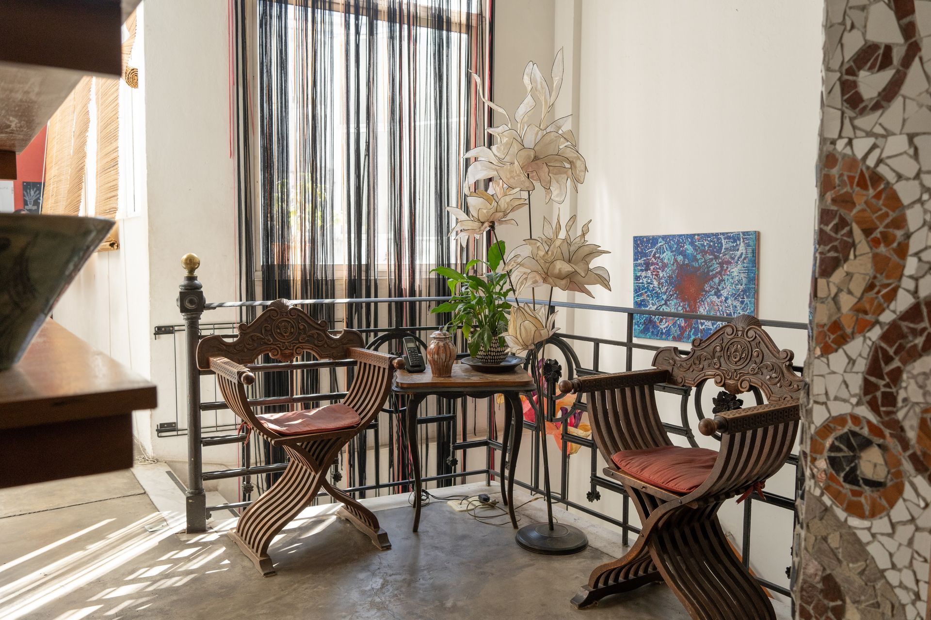 Balcón con sillas ornamentadas, mesa pequeña, cortinas transparentes y detalles de mosaico en la pared.