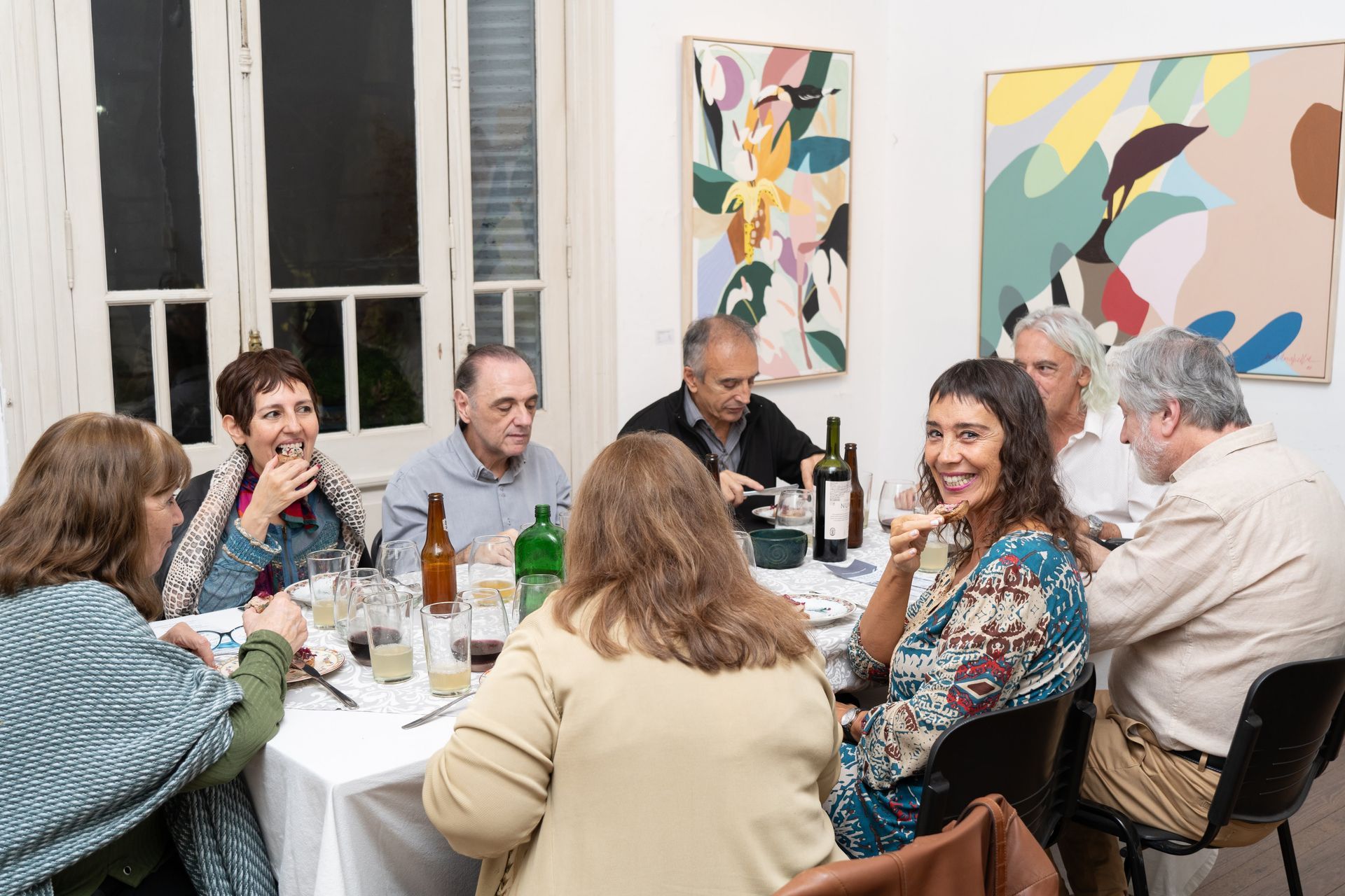 Personas sentadas alrededor de una mesa, cenando. Obras de arte en las paredes, botellas y vasos.