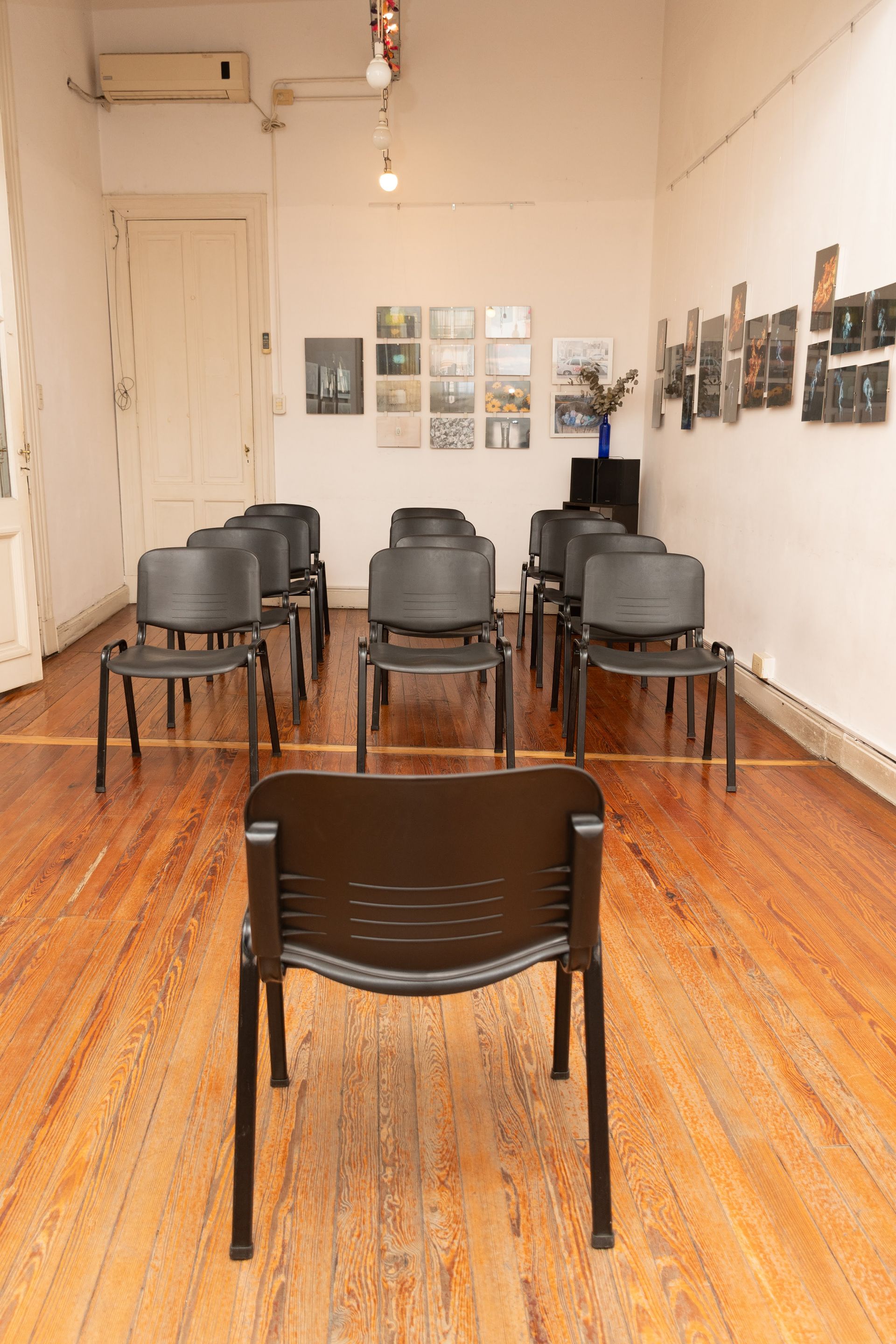 Una habitación vacía con pisos de madera, sillas negras dispuestas en filas y obras de arte en las paredes.