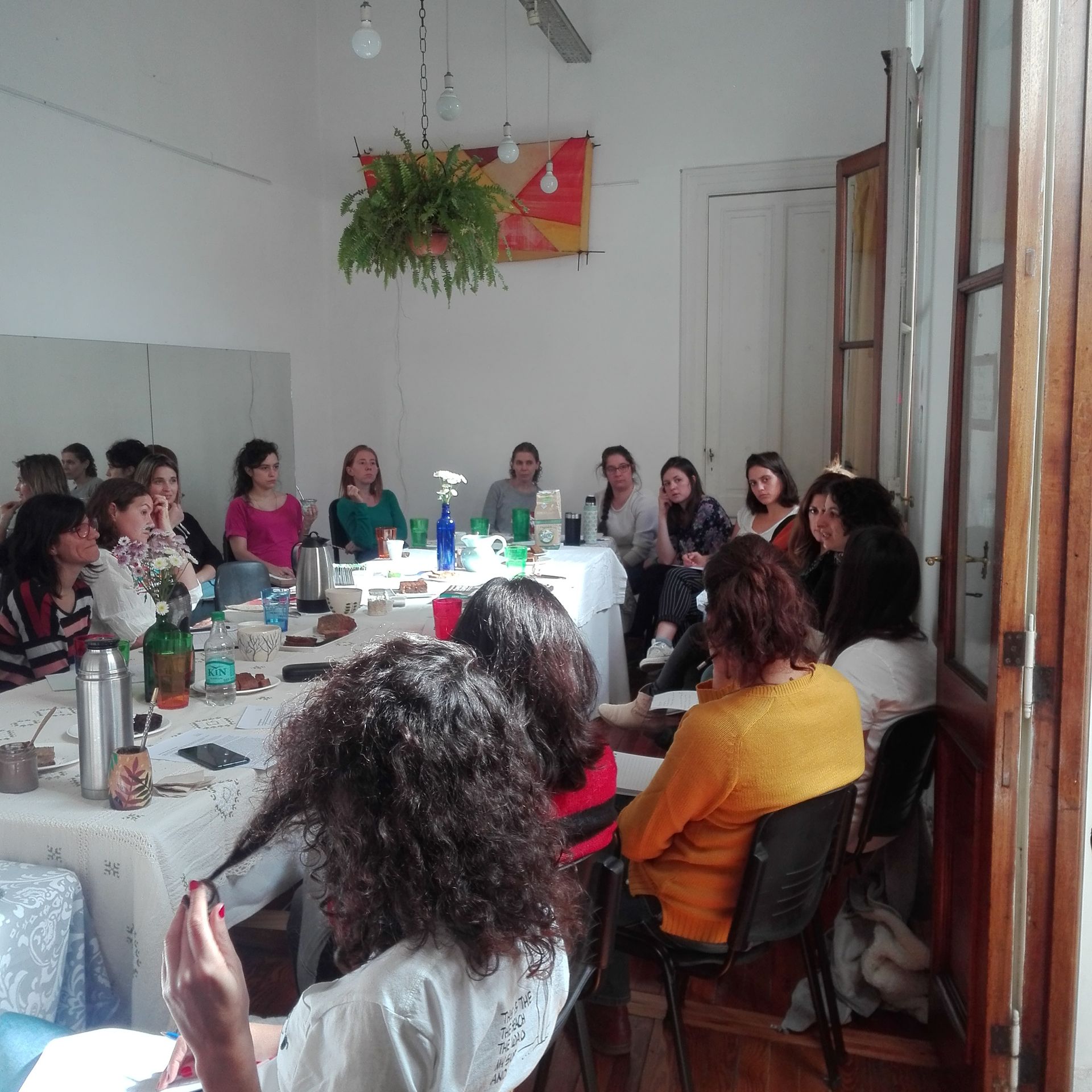 Personas sentadas alrededor de una mesa, asistiendo a una reunión en una sala con una planta y puertas abiertas.
