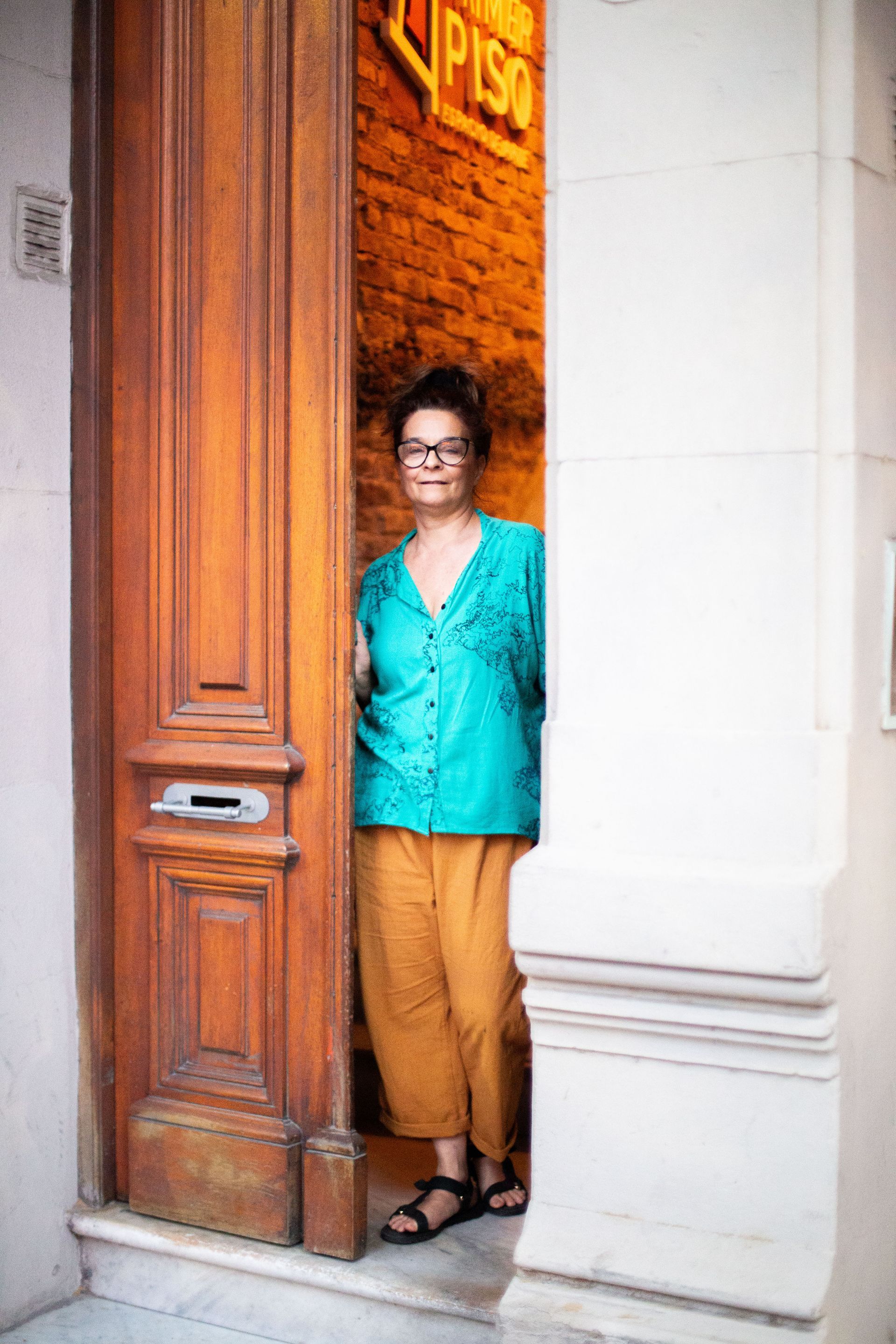 Una mujer con una camisa turquesa y pantalones amarillos está parada en la puerta.