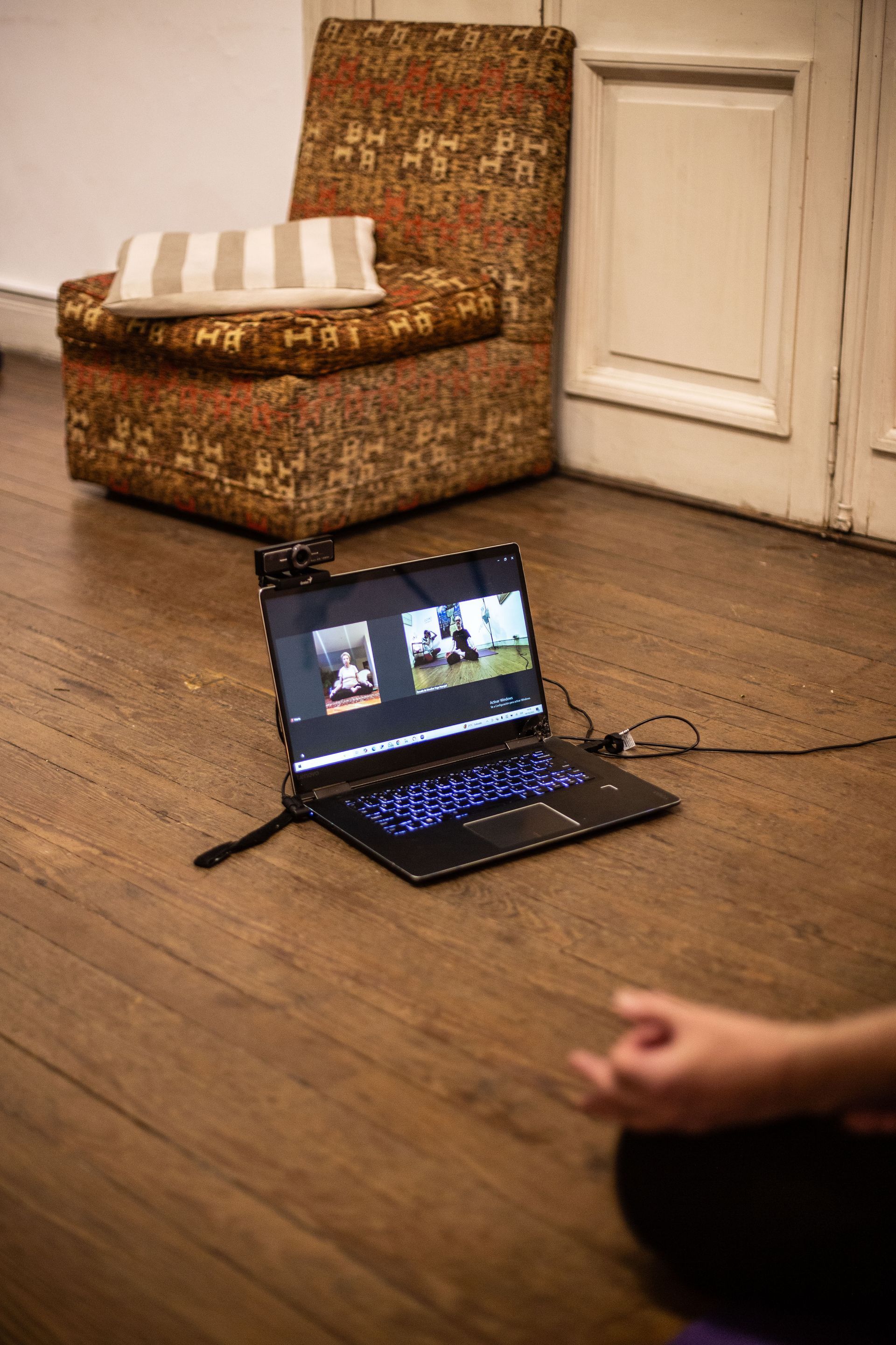 Portátil en el suelo con videollamada, cerca de una silla estampada. La mano de una persona en postura de meditación en primer plano.