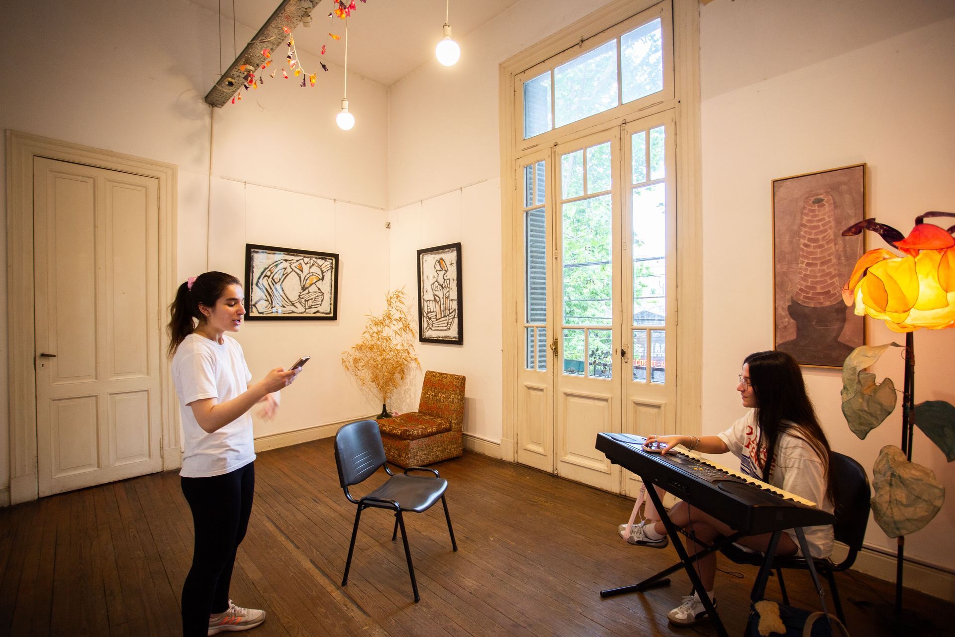 Dos mujeres en un estudio de arte: una con un teléfono, la otra tocando un teclado. Pinturas en las paredes, suelos de madera clara y ventanas luminosas.