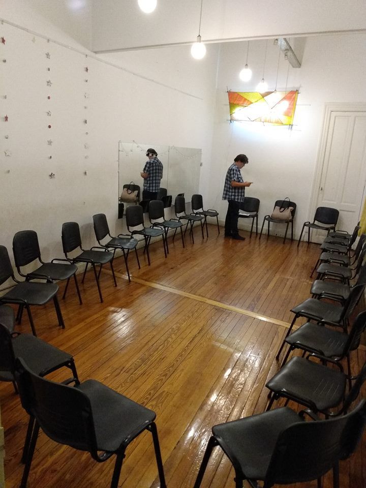 Una sala con sillas en círculo, con una persona de pie dentro. Suelo de madera, paredes blancas.