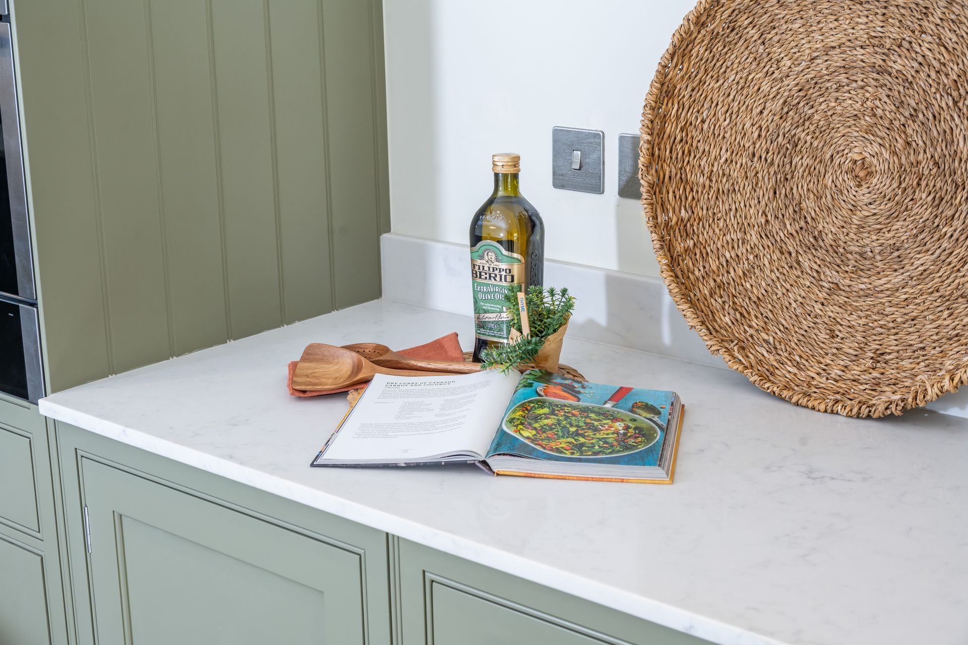 A kitchen counter with a bottle of olive oil , a cookbook , and a wicker basket.