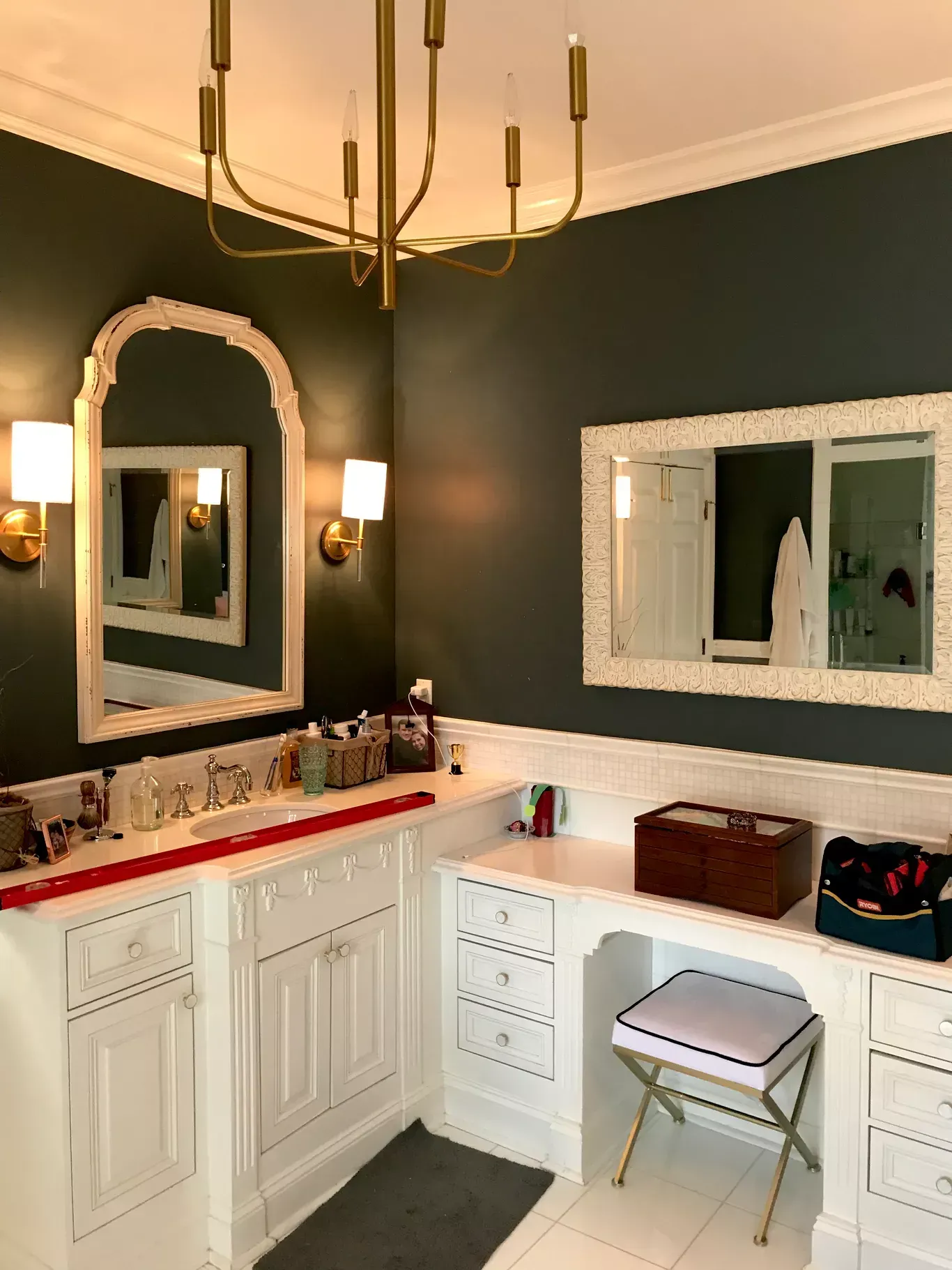 A bathroom featuring dark green walls, white cabinets, gold light fixtures, and two mirrors with white ornate frames.
