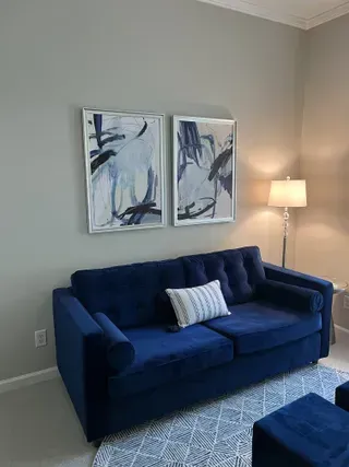 A navy blue sofa sits against a light wall, centered beneath two framed abstract blue-and-white art prints with a lamp.