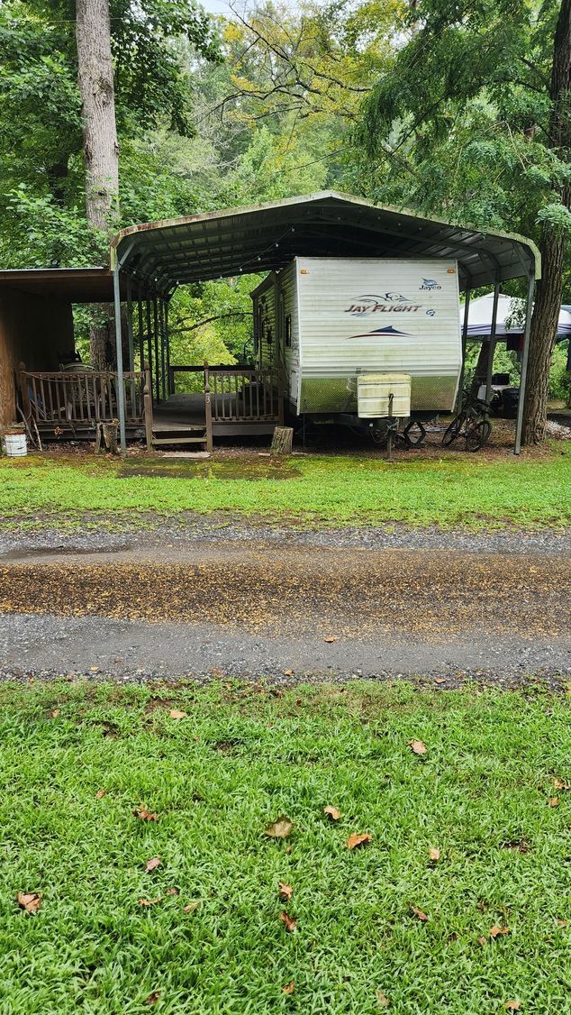 A white travel trailer under a metal carport in a wooded area with a gravel driveway.
