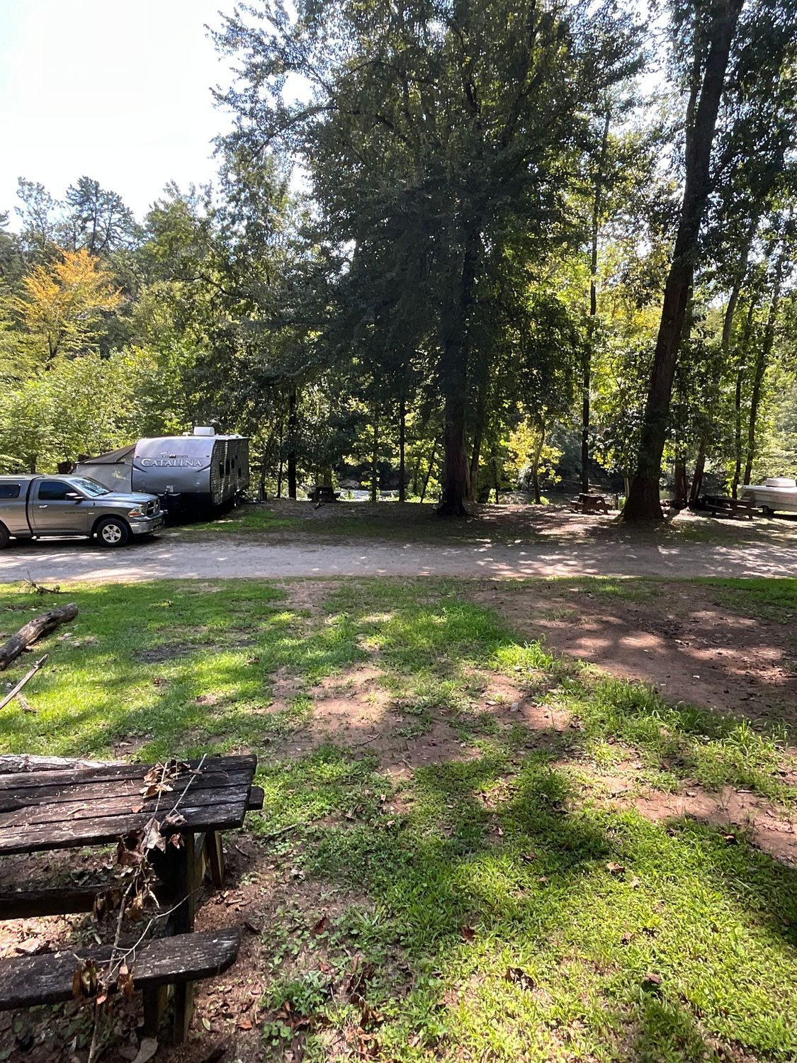 Camping scene: Vehicles and trailers in a wooded area with a picnic table in the foreground.
