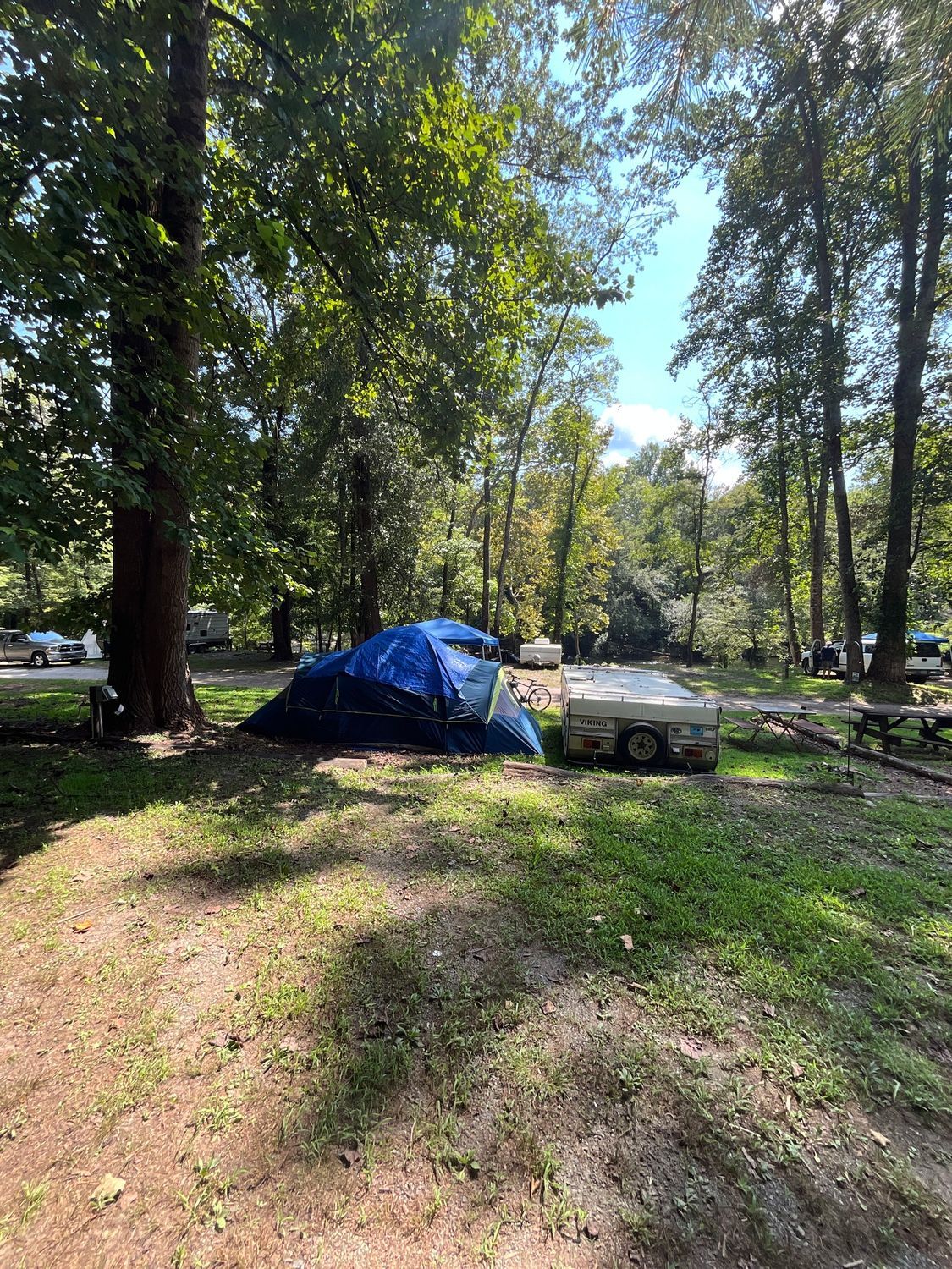 A blue tent and white trailer sit in a grassy campsite, surrounded by trees under a sunny sky.