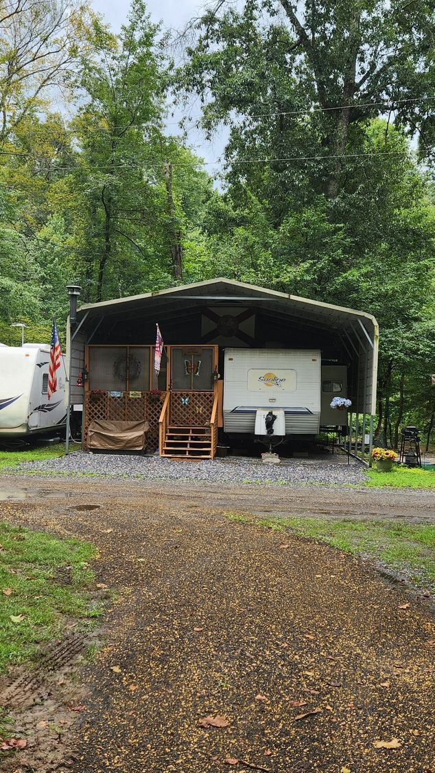 RV trailer under metal shelter with wooden steps, in a wooded area.