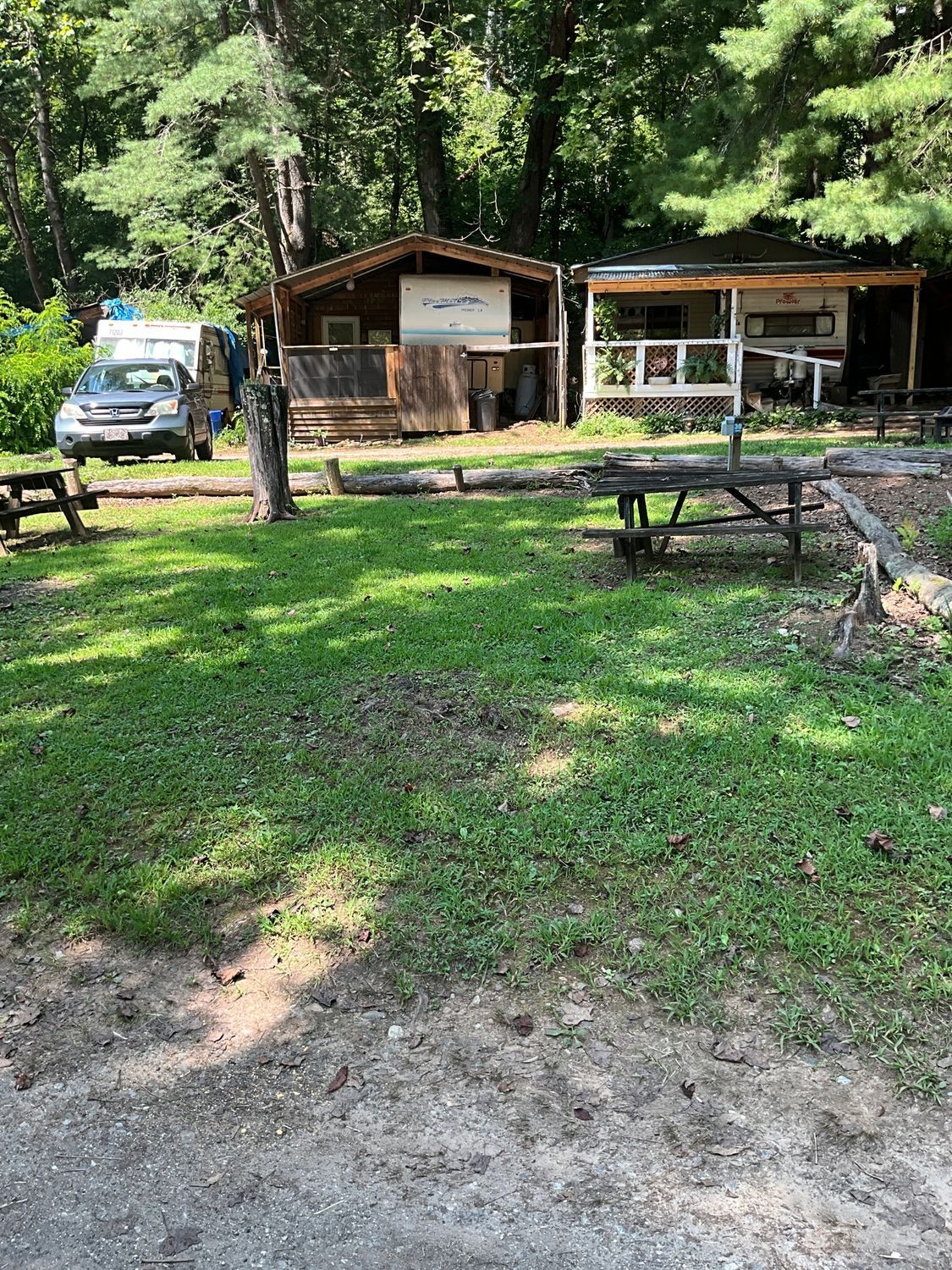 Two cabins with picnic tables and a vehicle on a grassy area with trees.