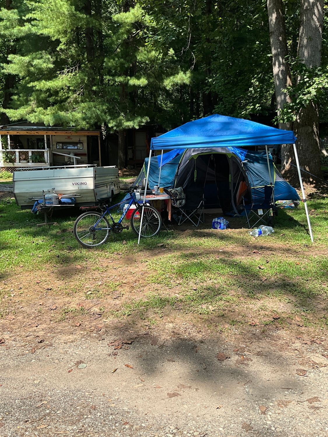 A campsite: blue tent under a canopy, bike, trailer, small building, trees.