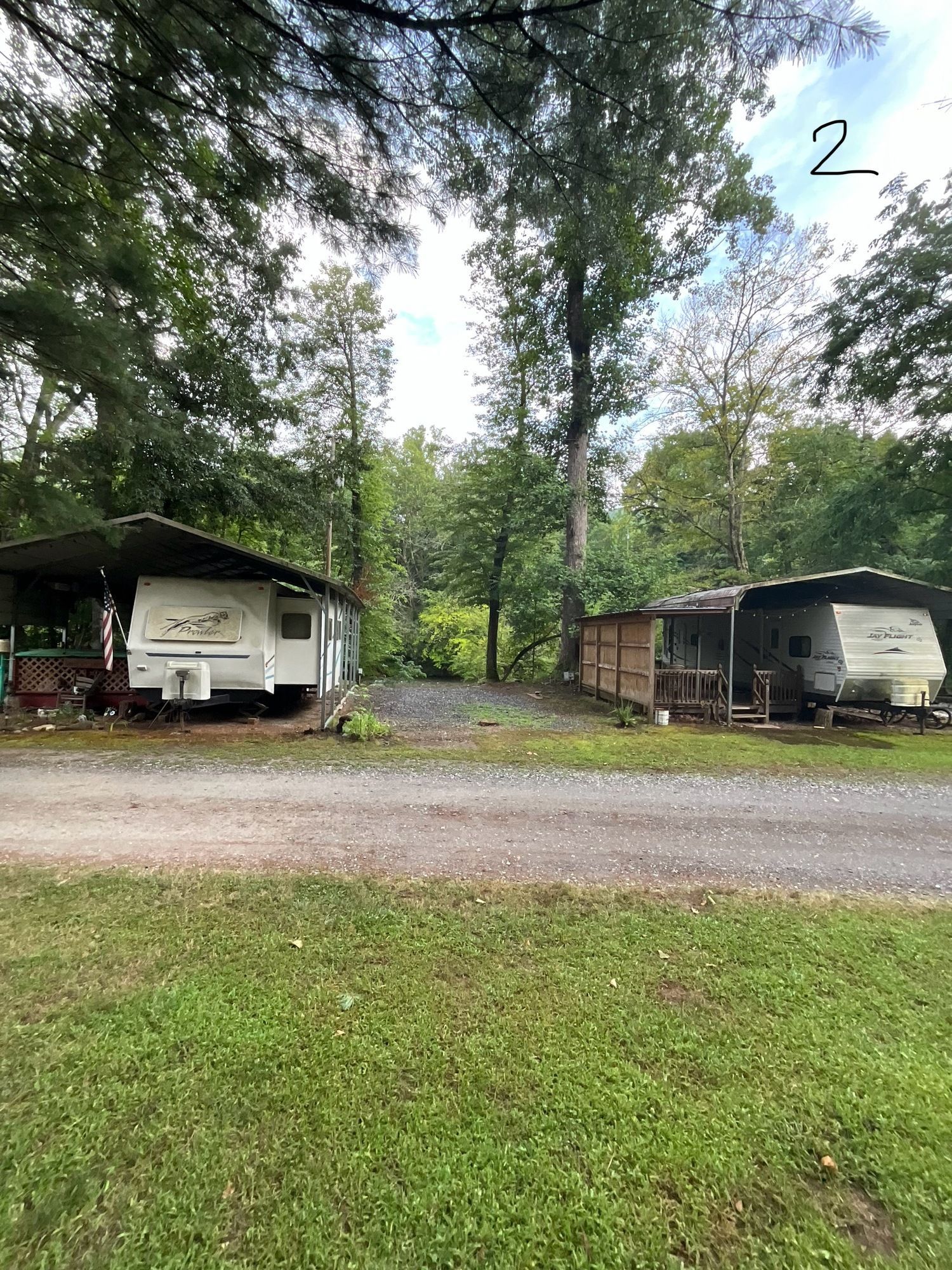 Two RV campers under covered shelters in a grassy, wooded area. Gravel driveway.