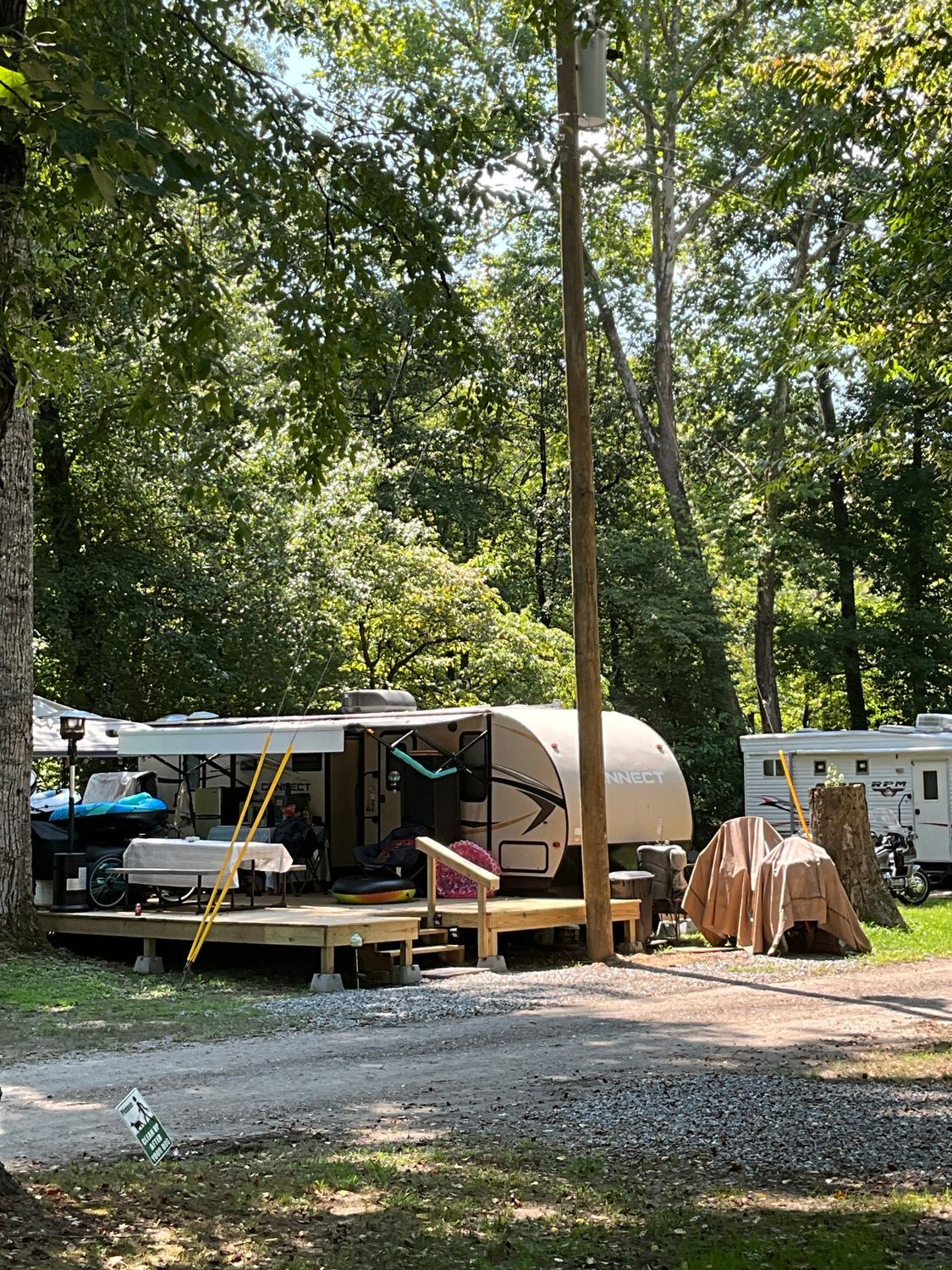 RV parked on a wooden deck in a wooded campground. Brown RV, green trees, and utility pole.