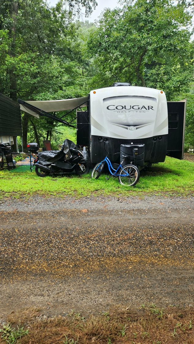 RV trailer parked on gravel with a recumbent bike and motorcycle nearby; surrounded by trees.
