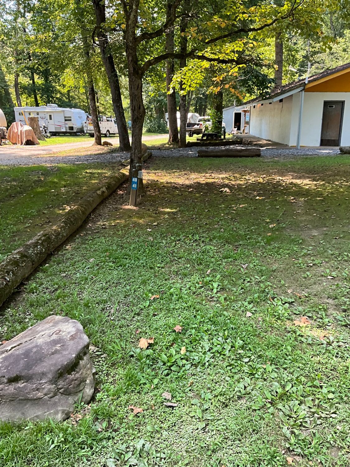 Grassy outdoor scene with trees, a log, a small building, and campers visible in the background.