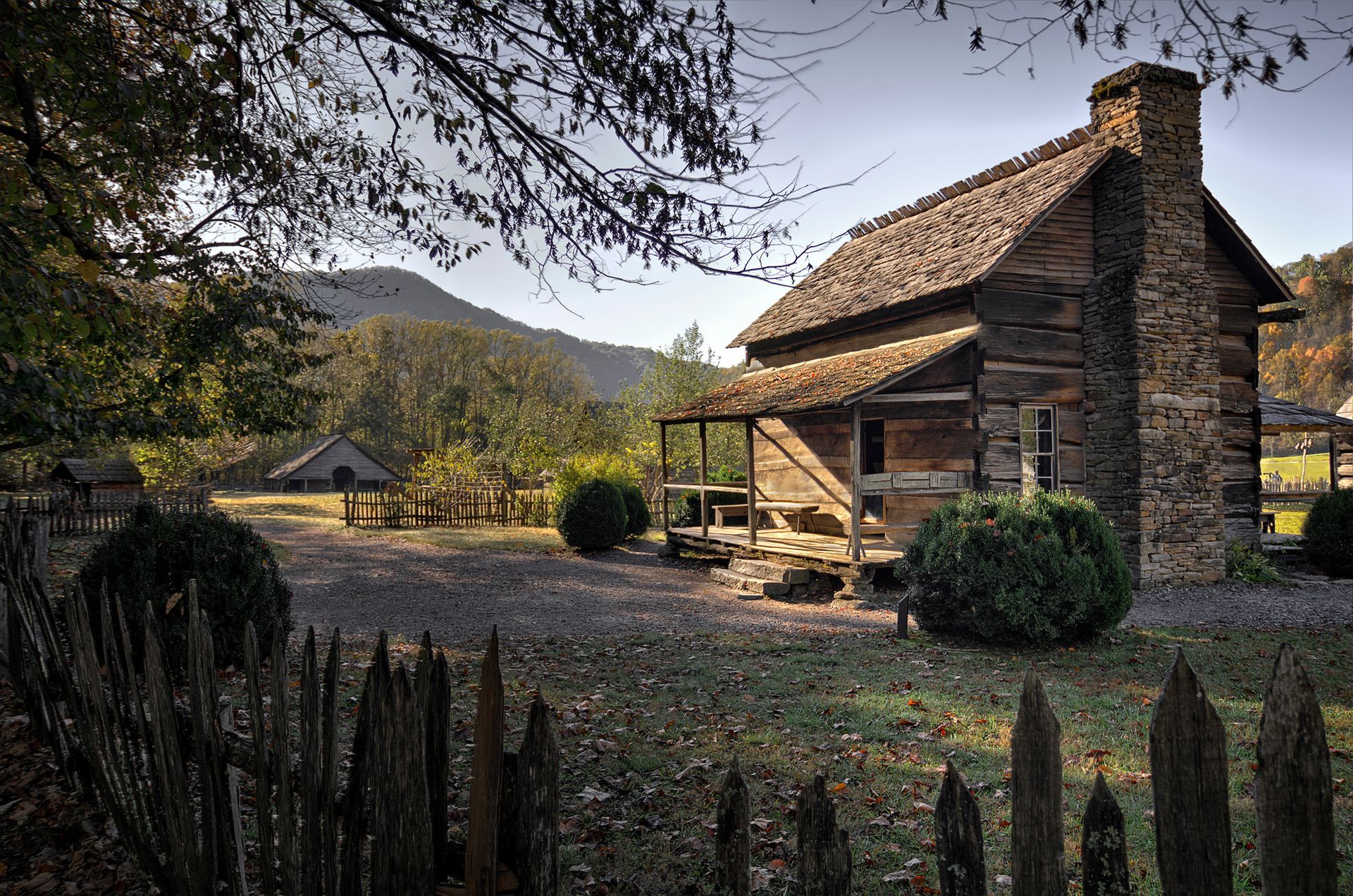 Rustic log cabin with stone chimney, surrounded by a wooden fence, trees, and hills in the background.