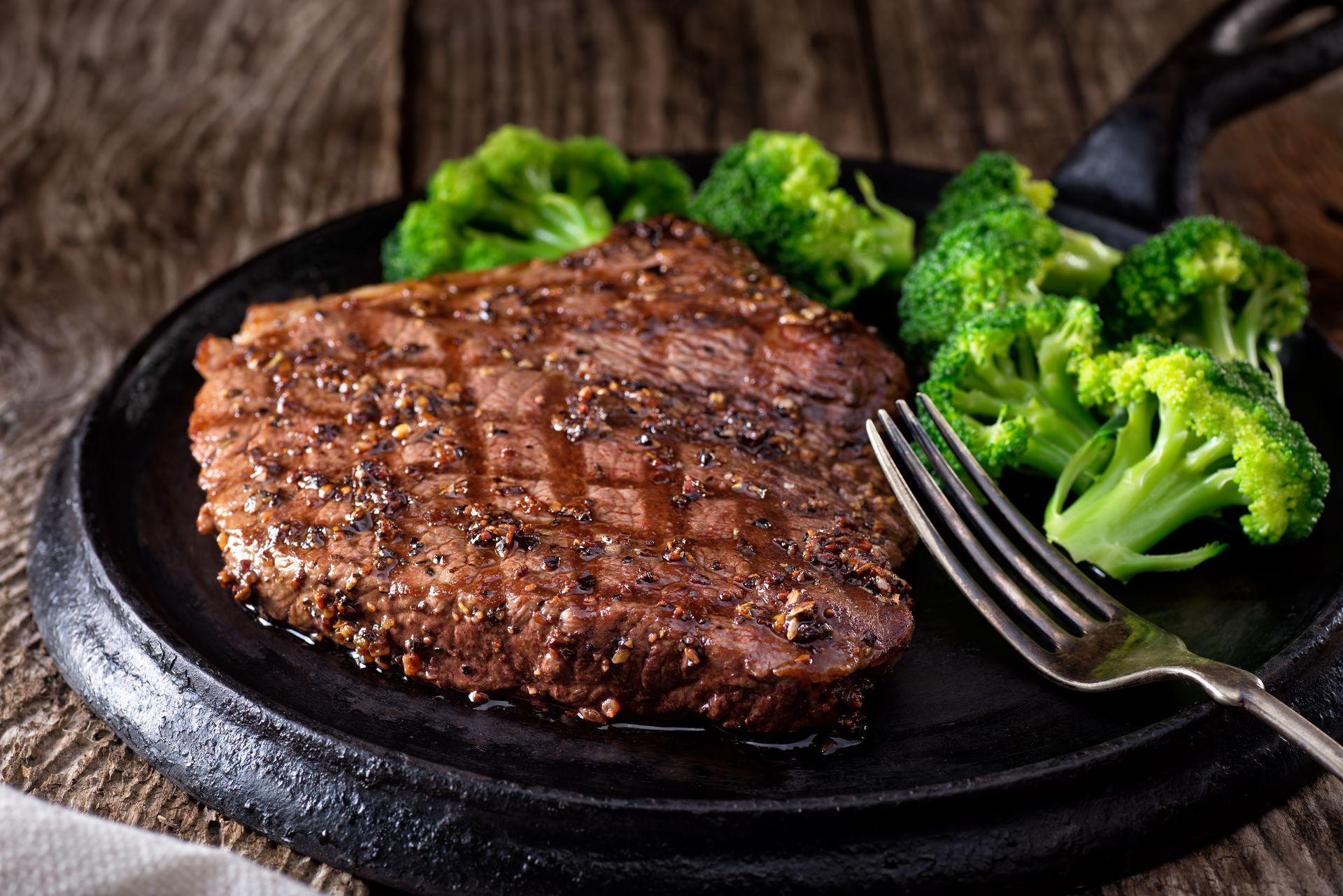 Grilled steak with broccoli on a black plate, with a fork on a wooden surface.