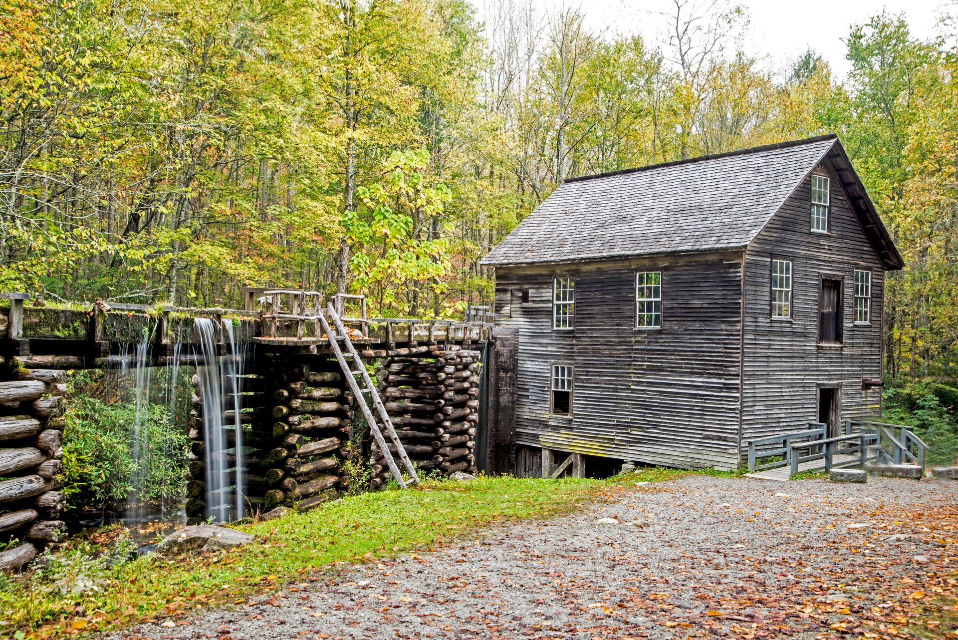 Old wooden mill with water flowing, surrounded by trees and a gravel path.