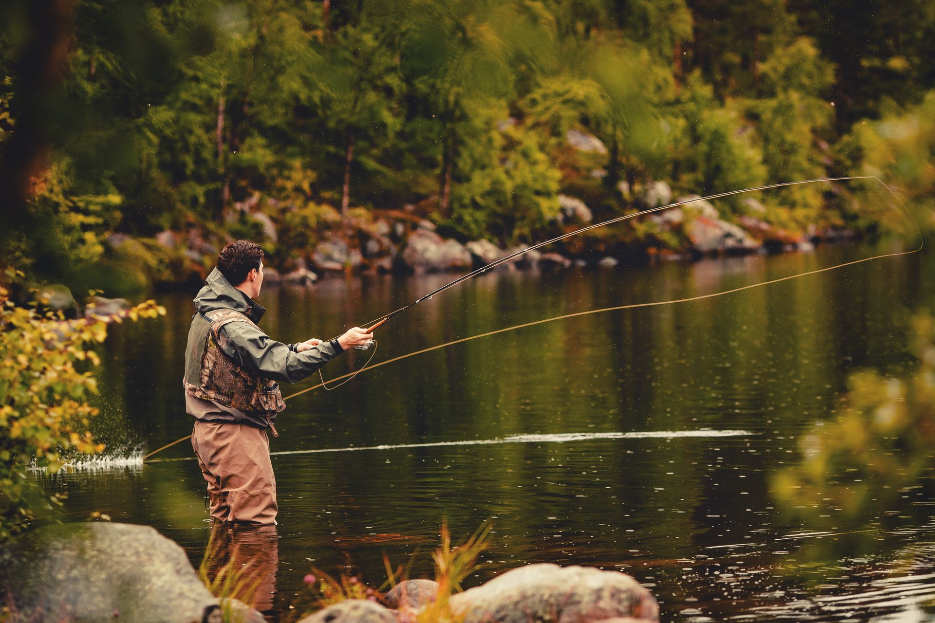 Man fly fishing in a calm river, wearing waders and vest; trees in the background.