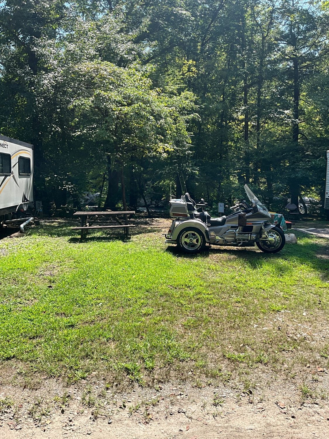 A trike motorcycle parked in a grassy campground next to a picnic table and RV in a wooded area.