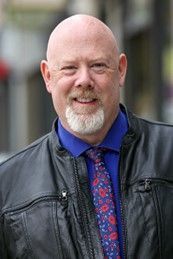 A bald man wearing glasses and a blue shirt is smiling for the camera.