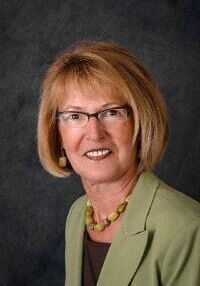 A woman wearing glasses and a green jacket is smiling for the camera.