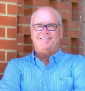 A man wearing glasses and a blue shirt is standing in front of a brick wall.