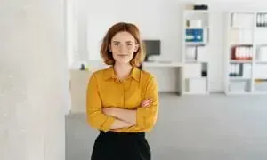 A woman in a yellow shirt and black pants is standing in an office with her arms crossed.