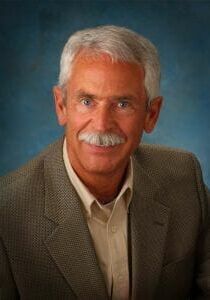 A man with gray hair and a mustache is wearing a suit and smiling for the camera.