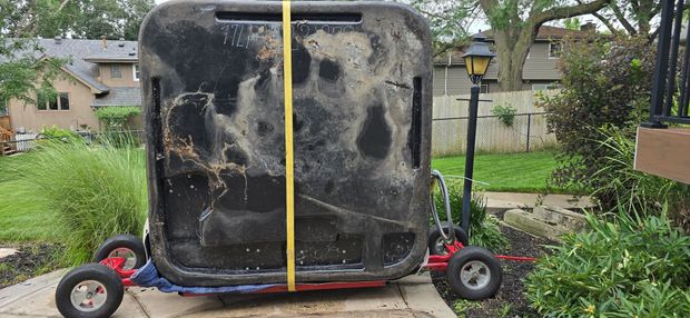 Large black appliance strapped upright on a small trailer in a garden, with red wheels and a yellow tie-down strap