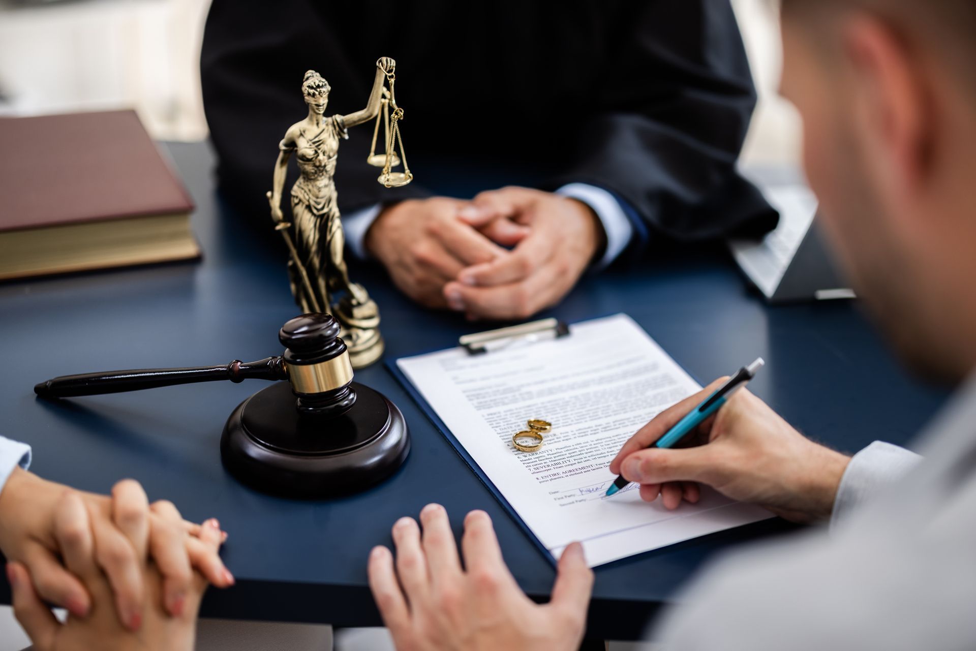 Man signing divorce papers in a courtroom with a judge and wedding rings visible on the document.