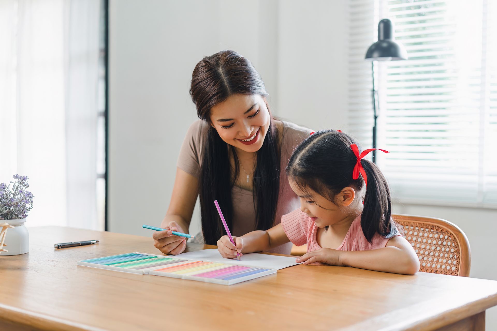 Woman and young girl drawing together at a table, indoors. The girl has a red bow.