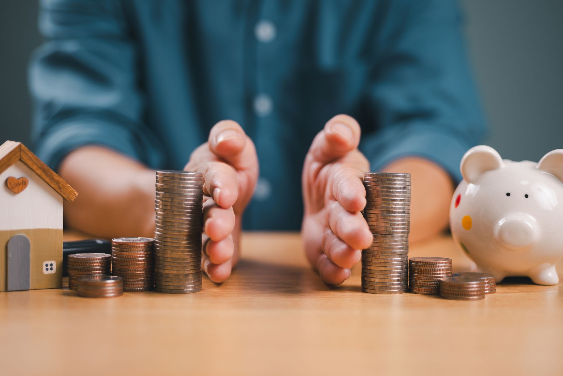 Hands protecting stacks of coins next to a house model and a piggy bank on a table.