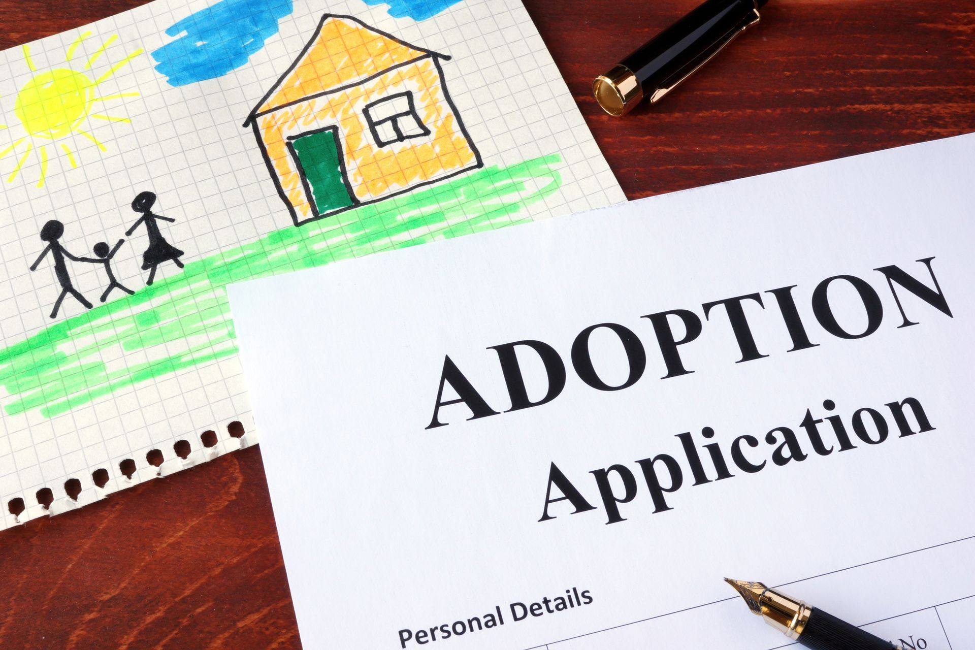 Adoption application form with a child's drawing of a family and house on a wooden table.
