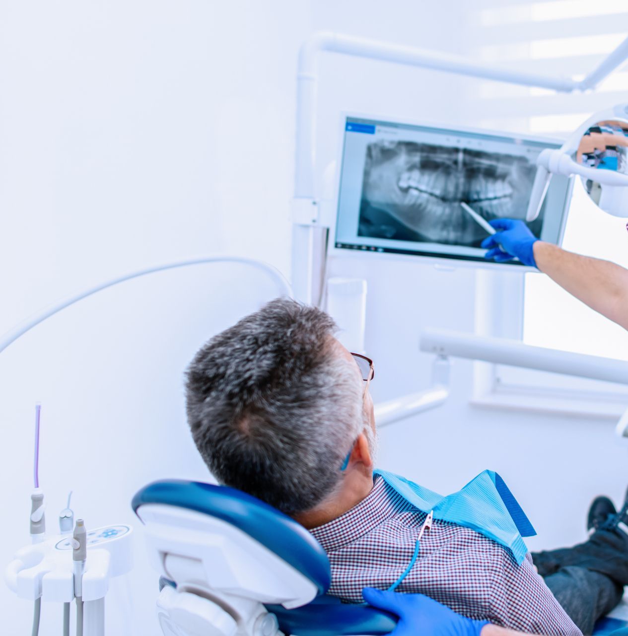 Dentista che mostra una radiografia dentale a un paziente. Stanza bianca. Guanti e bavaglino blu.
