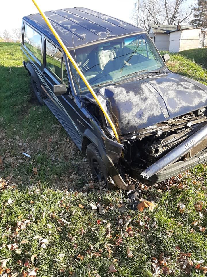 A black jeep is sitting in the grass with a yellow pole sticking out of the hood.
