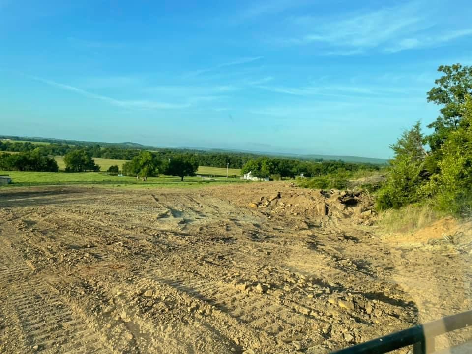 A wide open field of dirt and rock, ready for development, with green trees and clear blue sky in the background.