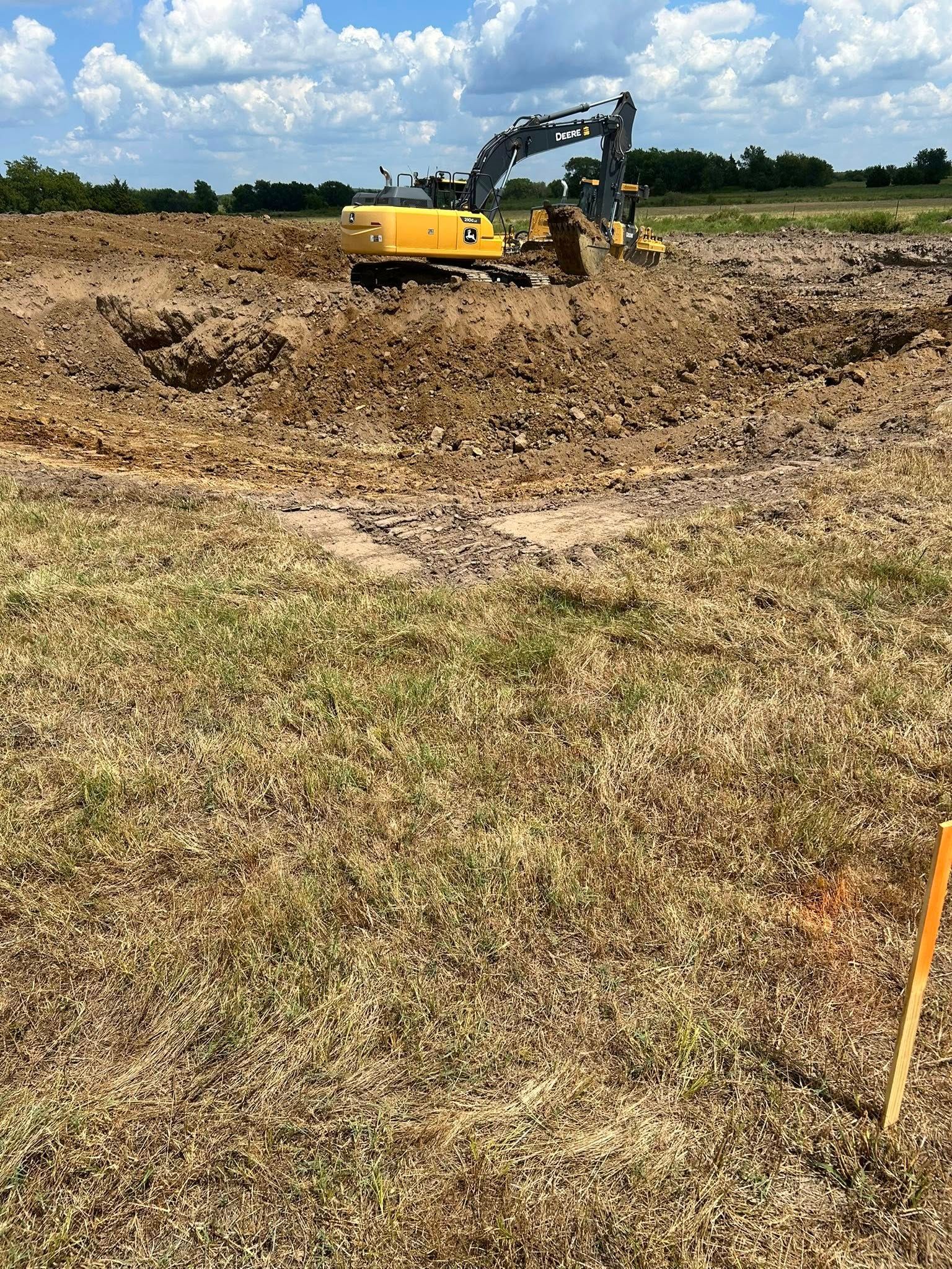 Yellow excavator digs in brown dirt, grassy field in foreground, blue sky background.