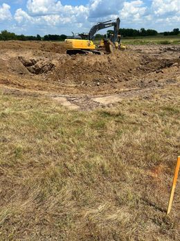 Yellow excavator digs in brown dirt, grassy field in foreground, blue sky background.