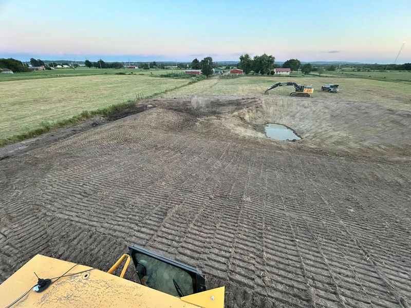 Heavy equipment grading a large, cleared field with a pond and structures in the distance.