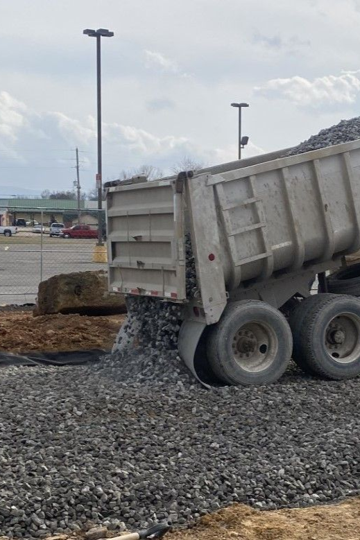 Dump truck dumping gravel onto a prepared surface in a parking lot. Gray rocks, cloudy sky.