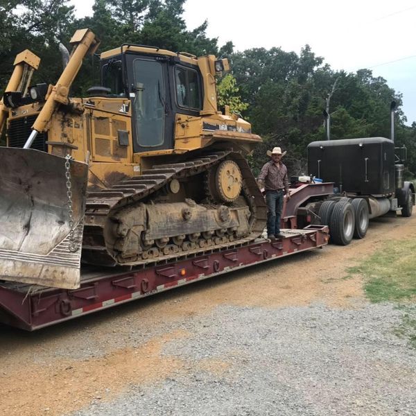 A man stands on a flatbed trailer carrying a yellow bulldozer, parked near a truck on a dirt road.